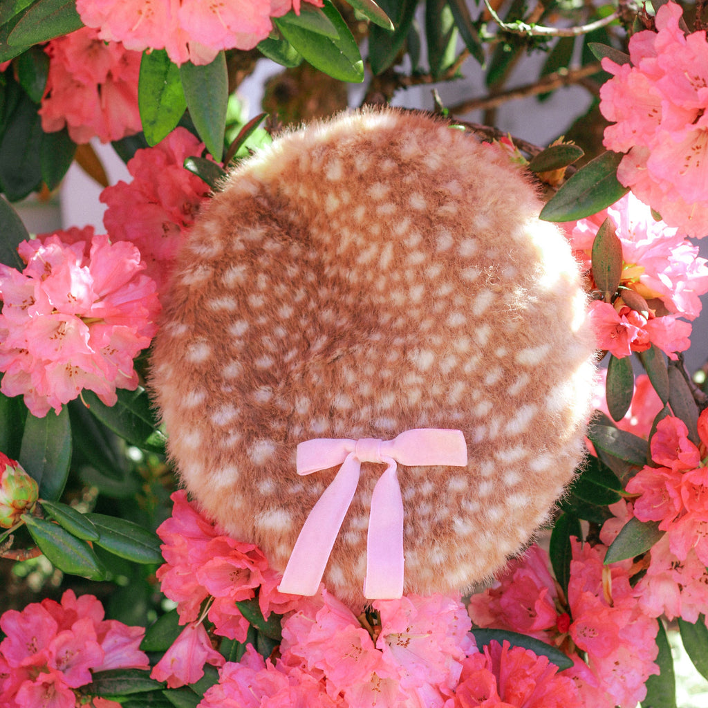 A round, fuzzy brown and cream spotted accessory with a light pink ribbon tied at the bottom, photographed outdoors among bright pink flowers and green leaves.